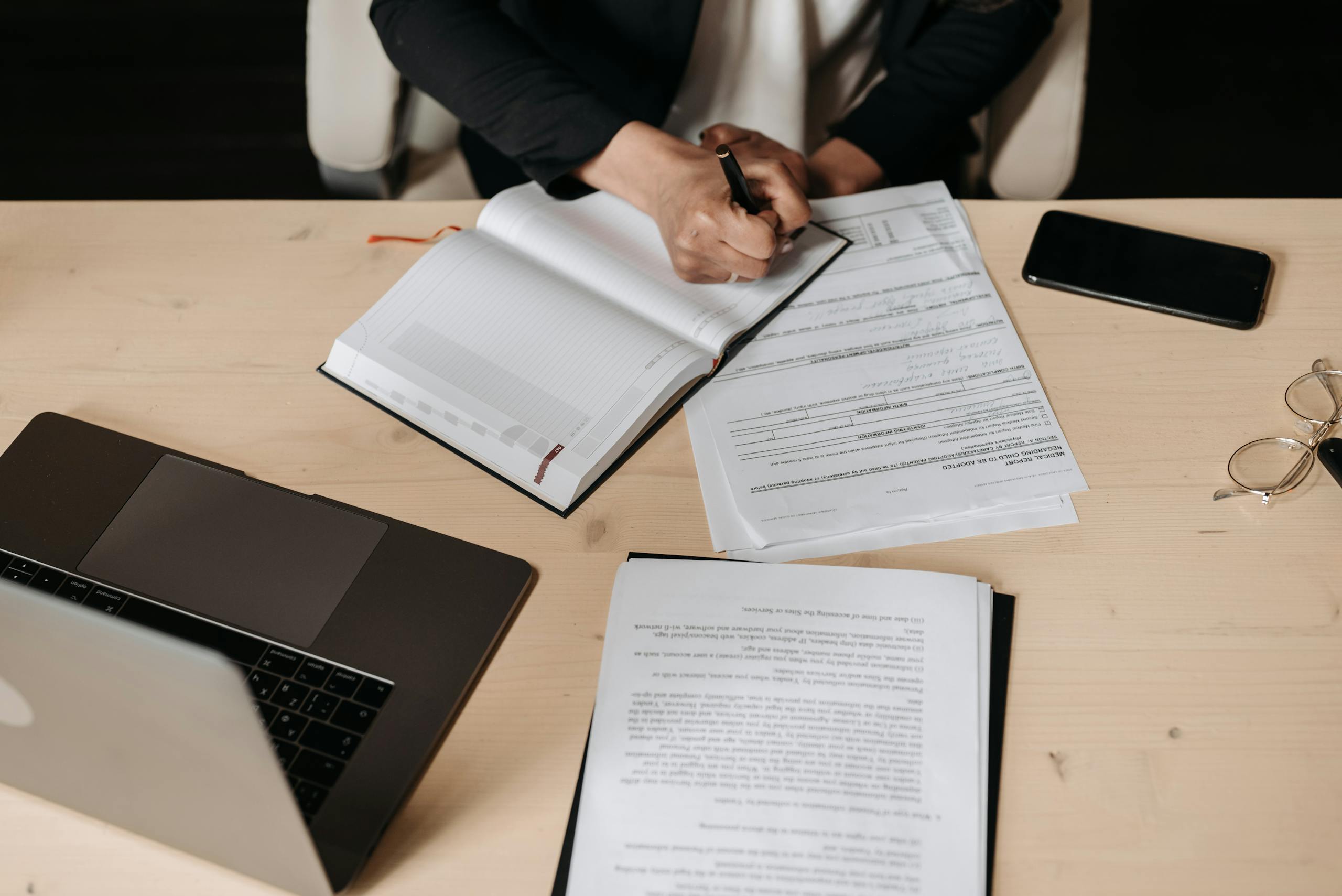 Overhead view of a desk setup with business documents, a laptop, and a person writing.